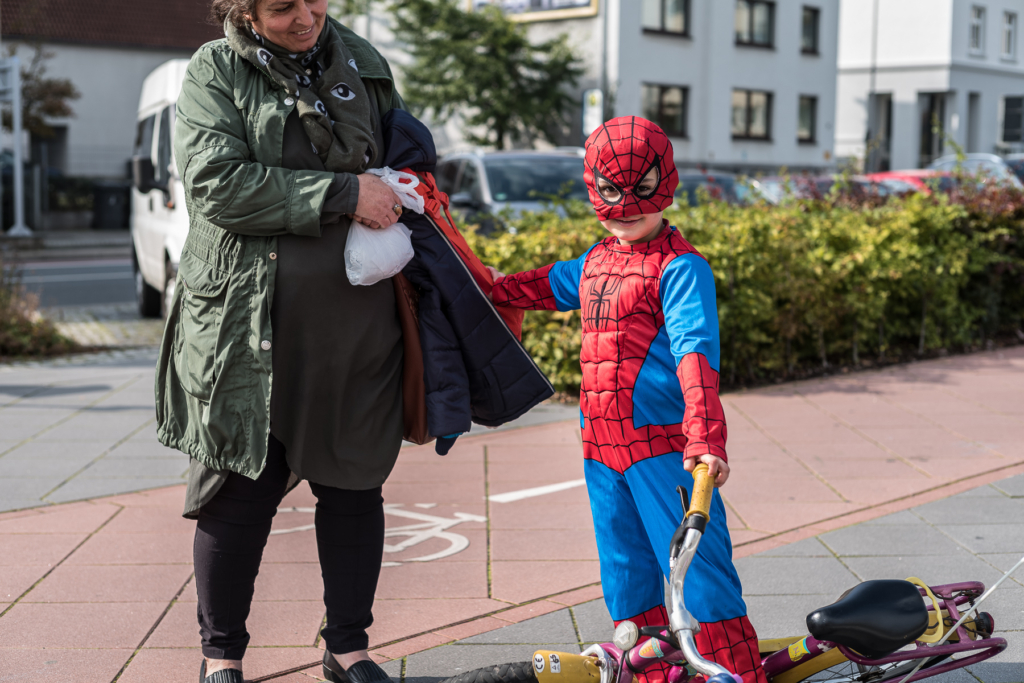Kind im Superheldenkostüm mit Fahrrad und Erwachsener auf Gehweg, geparkte Autos im Hintergrund.