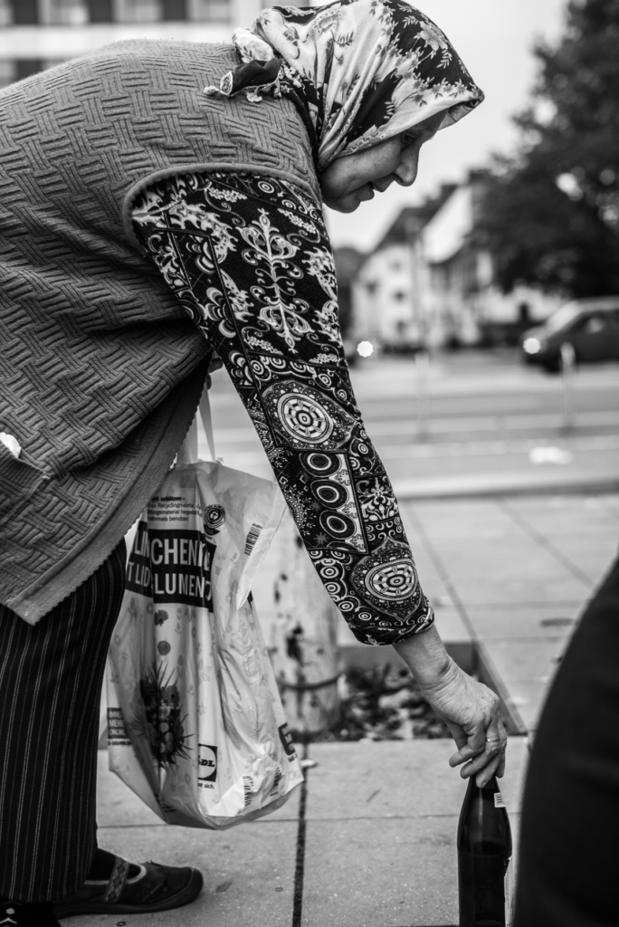 Ältere Frau mit Kopftuch hebt Flasche vom Boden, Einkaufstüte in Hand, städtischer Bürgersteig.
