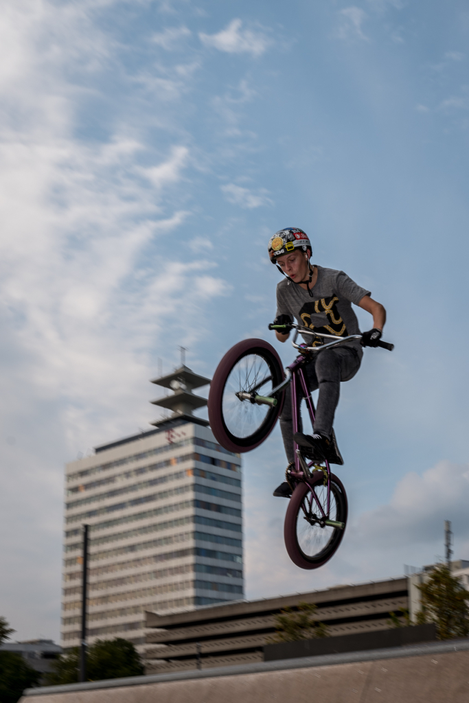 BMX-Fahrer in der Luft mit Helm vor modernem Gebäude und teilweise bewölktem Himmel