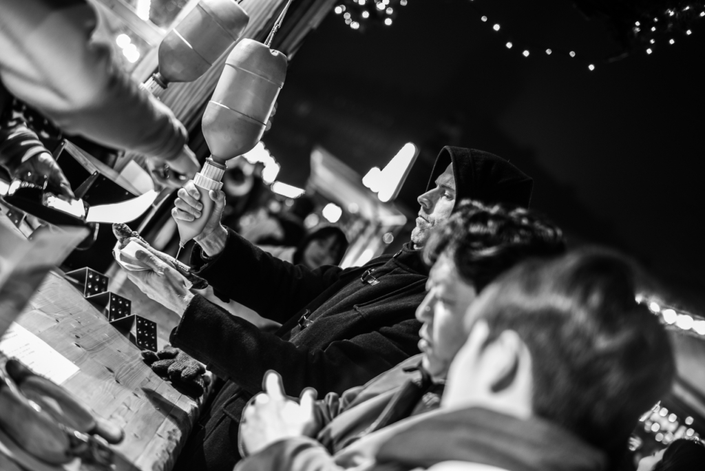 Belebter Markt bei Nacht, Person gibt Soße auf Gericht, Lichter im Hintergrund, Verkaufsstand.