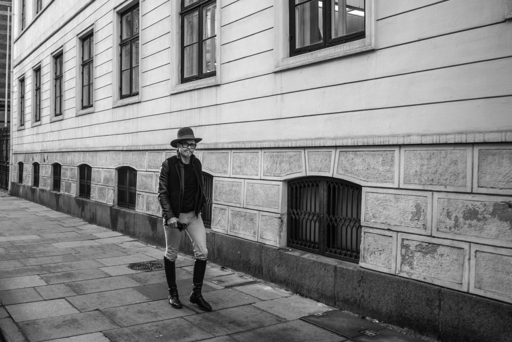 Person mit Hut und Brille geht auf Gehweg neben Gebäude mit großen Fenstern in Schwarzweiß.