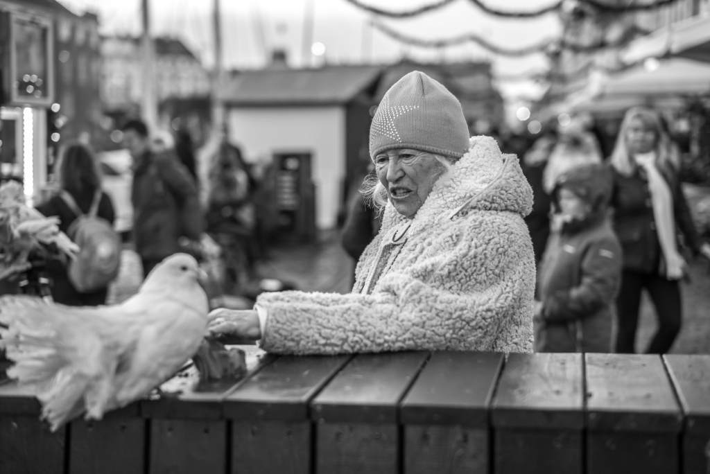 Ältere Person in flauschiger Jacke am Tisch mit Taube, unscharfe Personen im Hintergrund draußen.