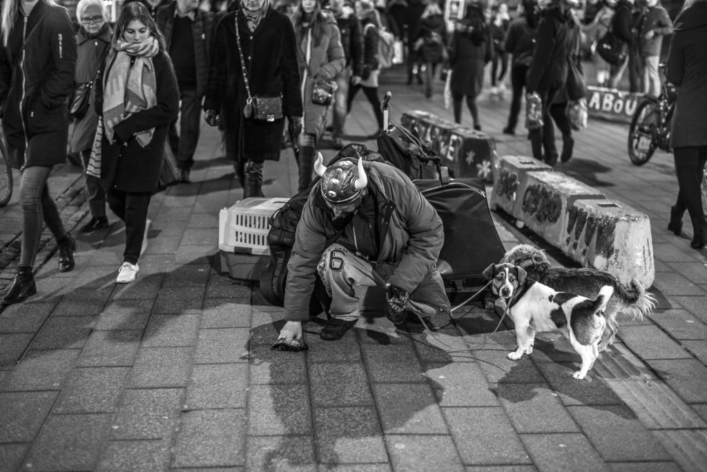 Person mit zwei Hunden hockt auf belebtem Bürgersteig, hebt etwas auf, Passanten gehen vorbei.