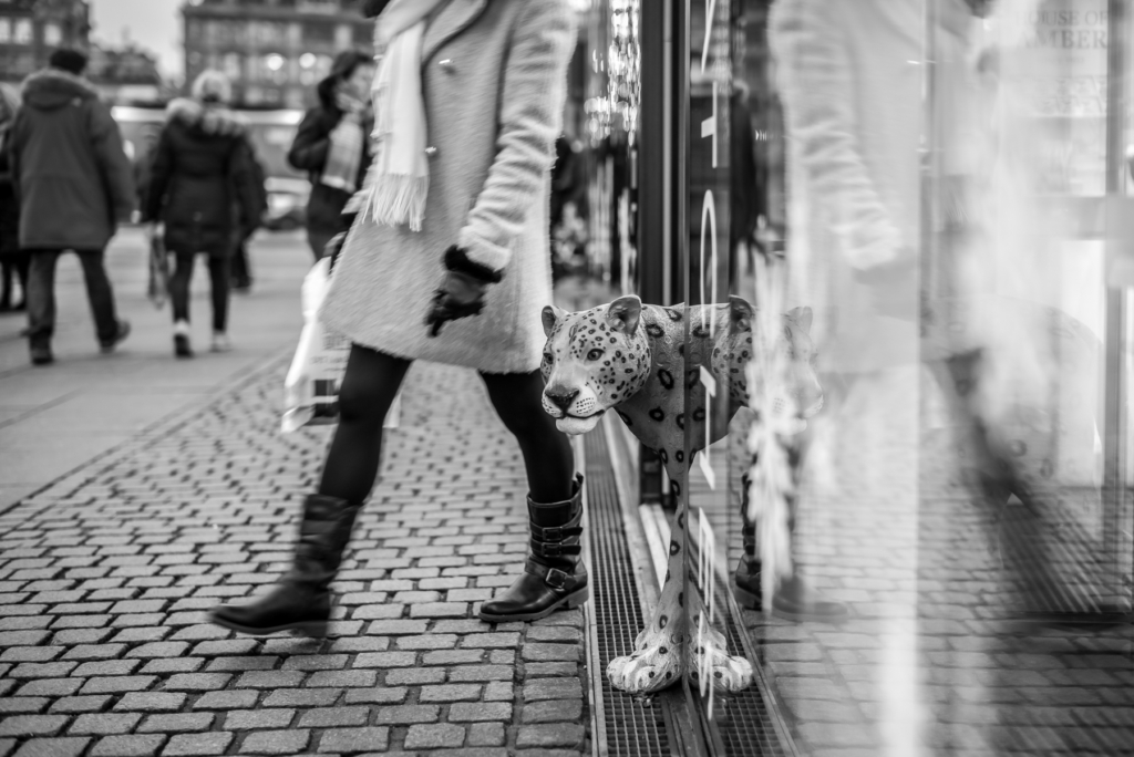 Belebte Straße mit Passanten, Person im Mantel vor Schaufenster, Skulptur eines gepunkteten Tieres.