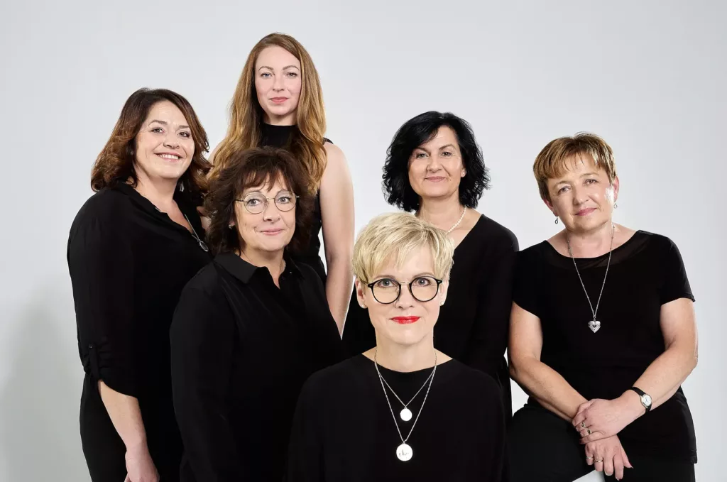 Sechs Frauen in schwarzer Kleidung vor neutralem Hintergrund, mit Schmuck und Brillen.