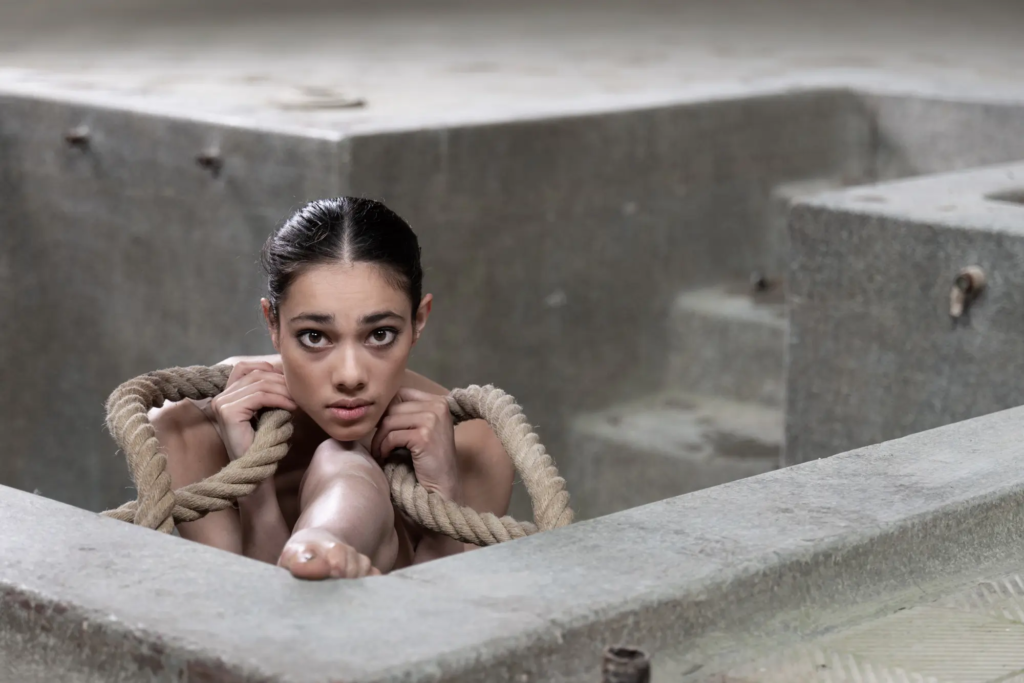 Frau mit Seil in leerer Betonstruktur sitzend, schaut direkt in die Kamera.