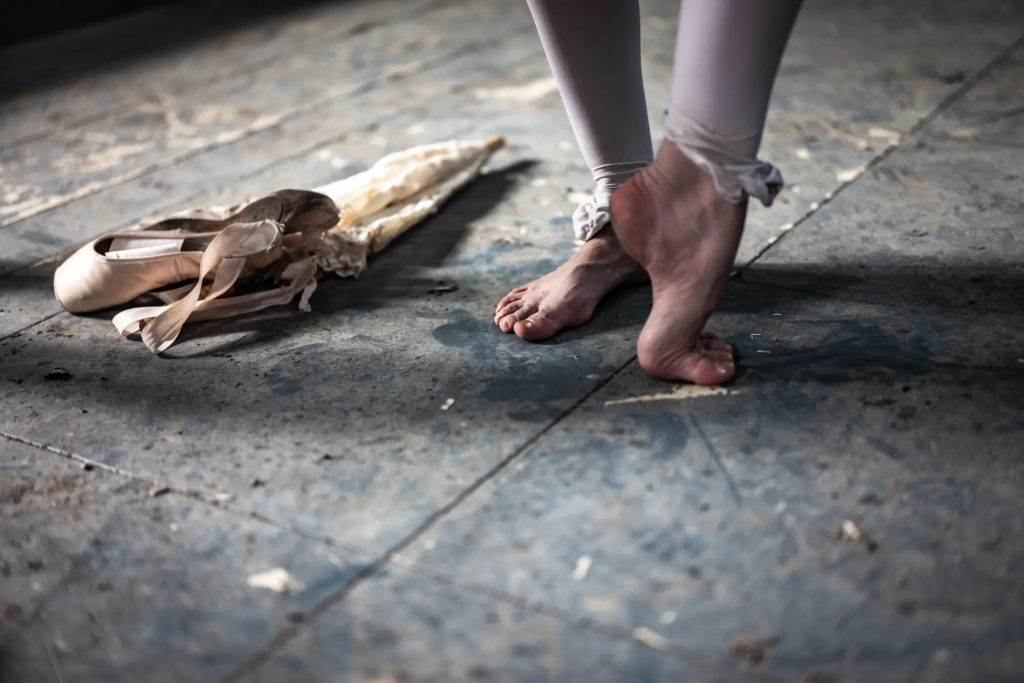Ballettschuhe auf abgenutztem Boden neben barfüßigen Füßen in rosa Strumpfhosen.