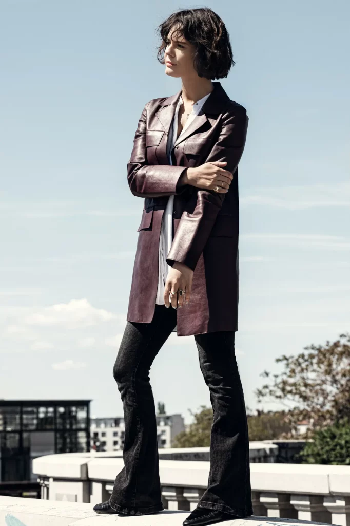 Person im Ledermantel und Schlaghosen steht draußen vor städtischem Gebäude und klarem Himmel.