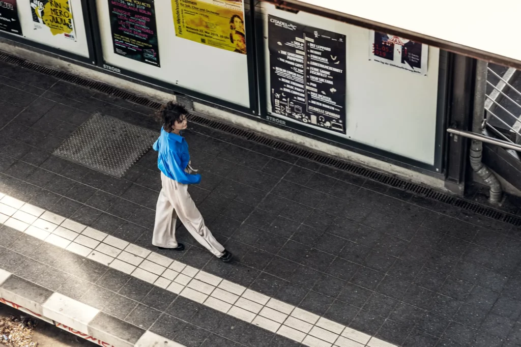 Person im blauen Hemd und weißen Hosen auf Bahnsteig, vorbei an Wandplakaten gehend.