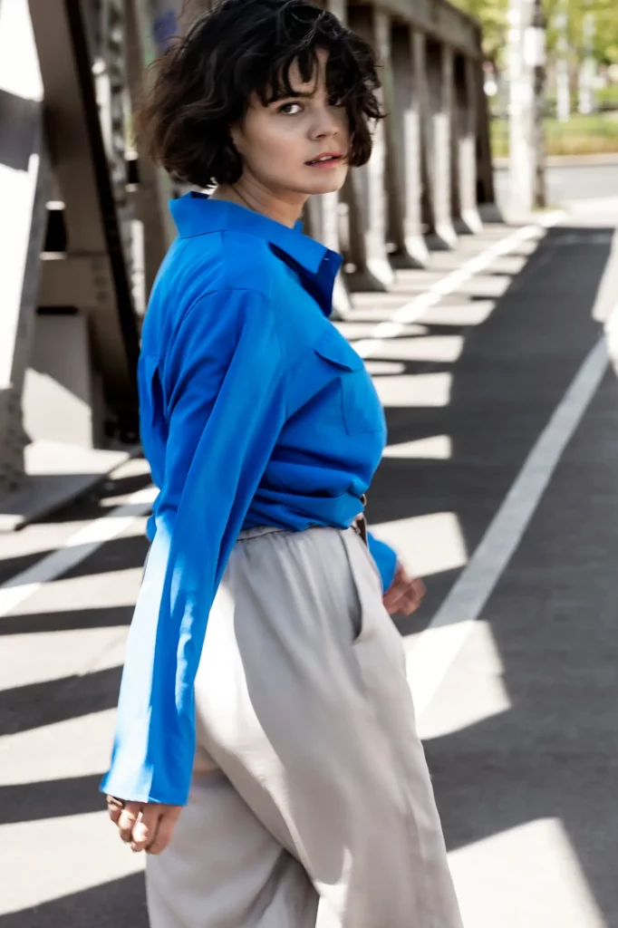 Frau in blauer Bluse und heller Hose auf Straße vor Brücke im Schatten.