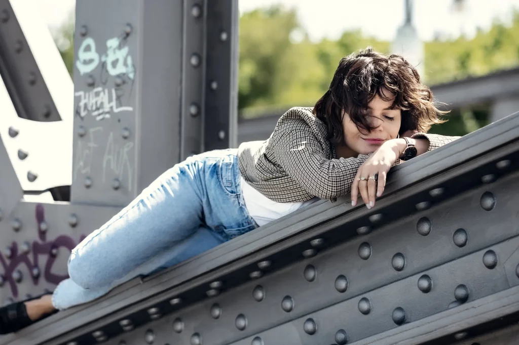 Person entspannt auf metallener Brücke mit Graffiti, trägt karierte Jacke und Jeans, Bäume im Hintergrund