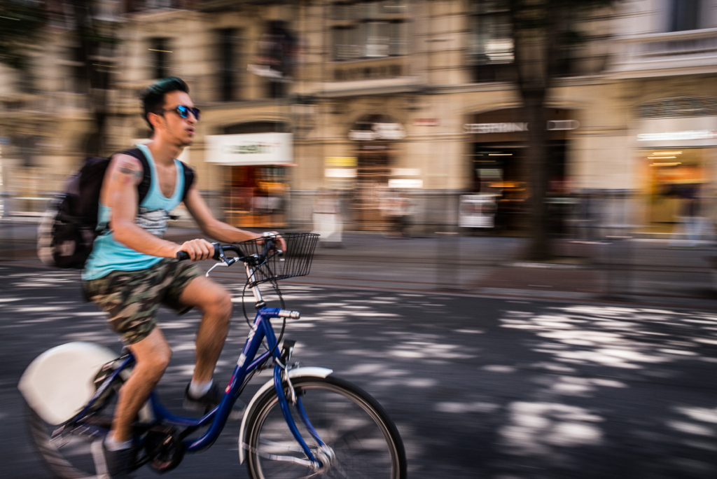 Person auf Fahrrad mit Bewegungsunschärfe in einer urbanen Straße, Gebäude im Hintergrund.