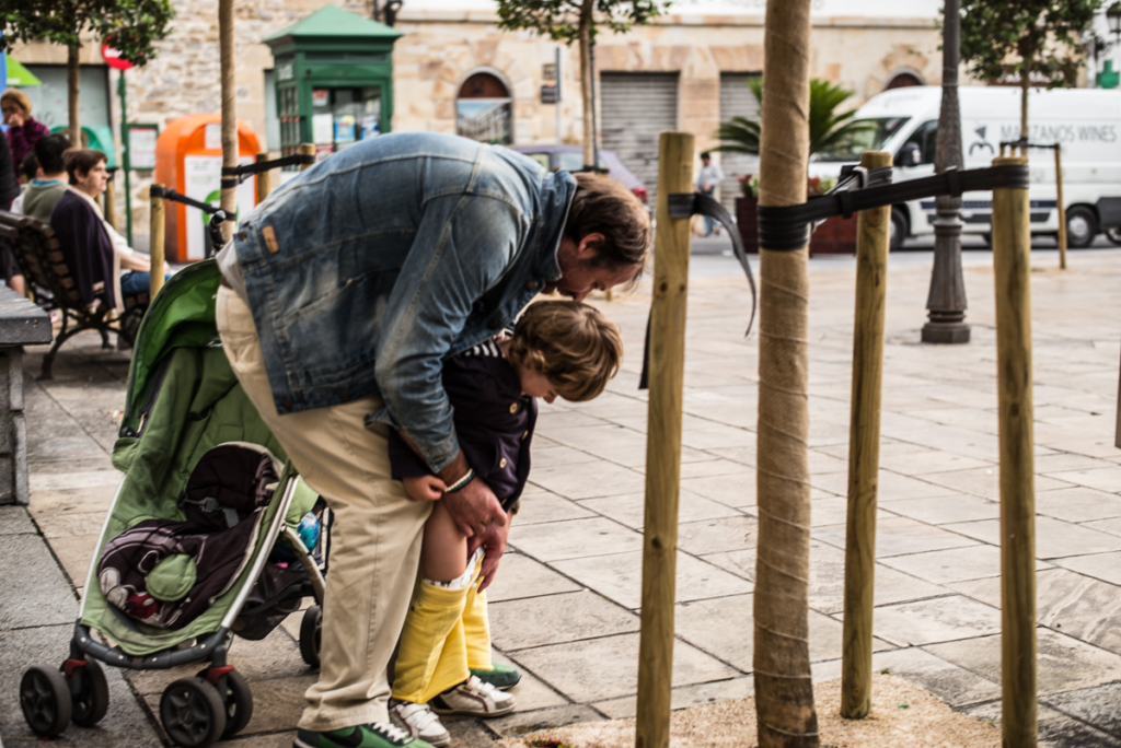 Ein Erwachsener hilft einem Kind, Hose hochziehen, mit Kinderwagen in städtischem Umfeld.