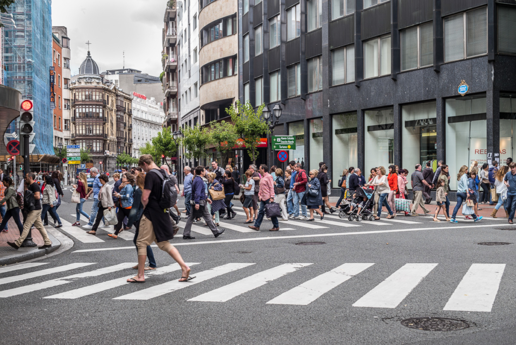 Große Menschenmenge überquert belebte Straße in Stadt mit modernen und historischen Gebäuden.