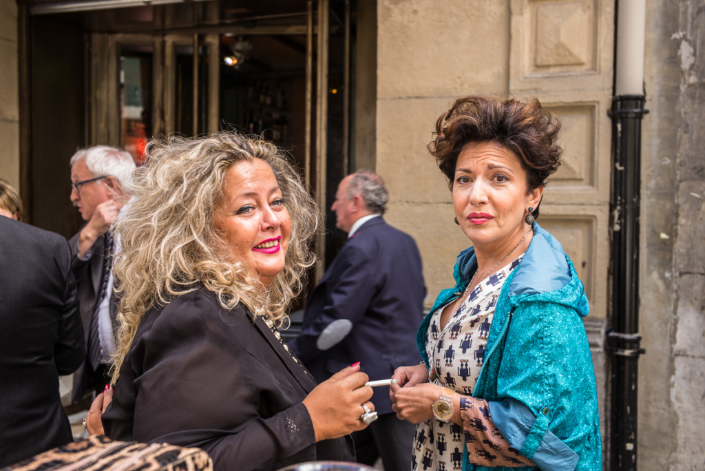 Zwei Frauen vor einem Eingang, blonde mit dunkler Kleidung, braune Haare mit türkisfarbener Jacke.