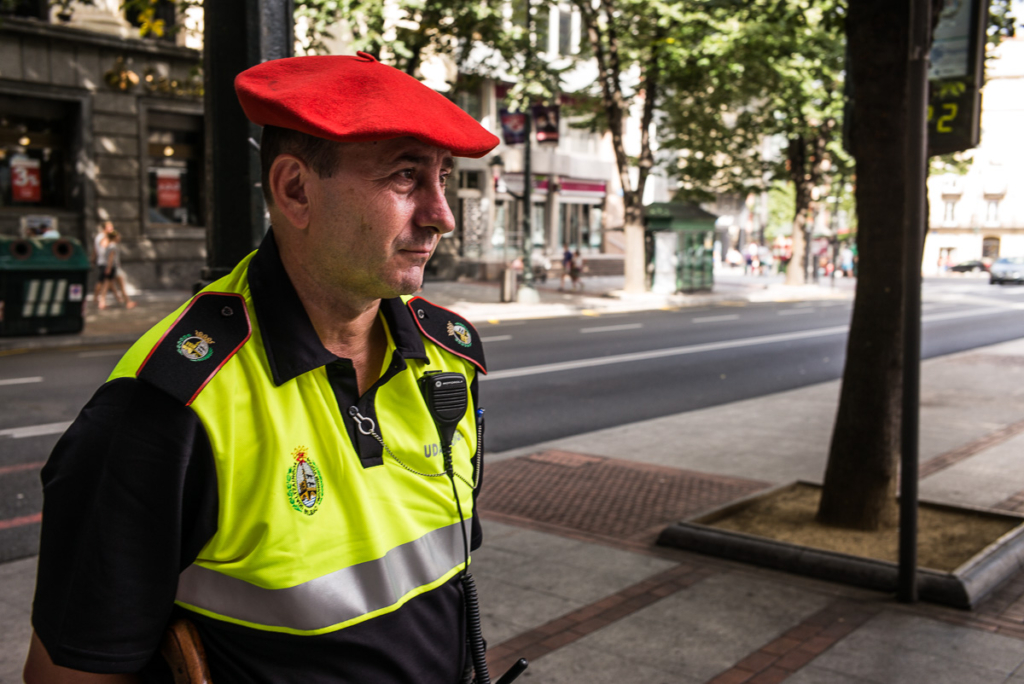 Uniformierter Mann mit rotem Barett auf städtischer Straße, Bäume und Gebäude im Hintergrund.
