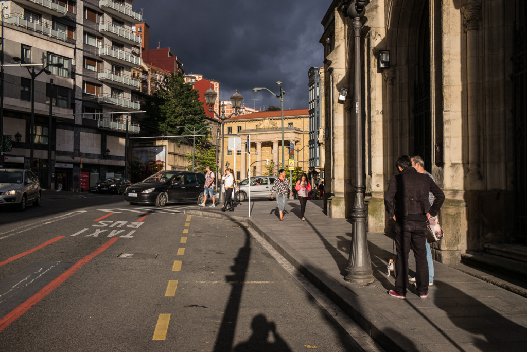 Städtische Straße bei bewölktem Wetter mit Passanten, Autos und Gebäuden im Hintergrund.