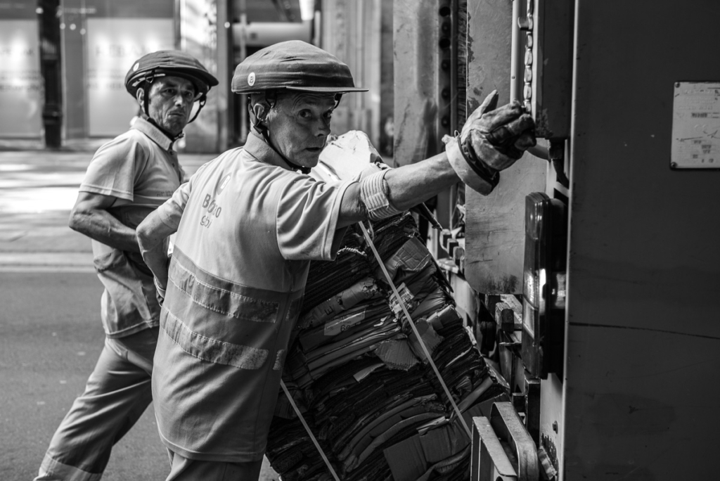 Zwei Arbeiter verladen Papier in ein Fahrzeug auf einer Straße, mit Helmen und Sicherheitskleidung.