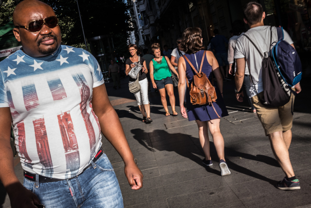 Menschen in Freizeitkleidung mit Rucksäcken auf sonniger städtischer Straße, Gebäude im Hintergrund.