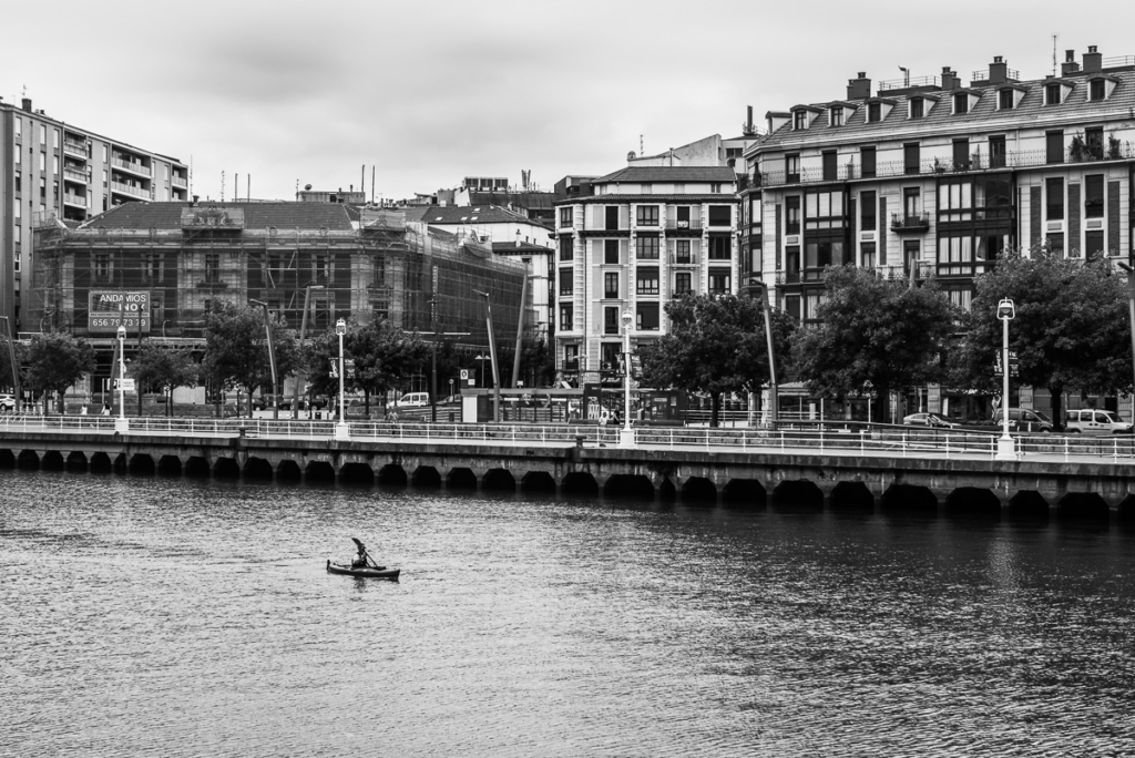 Städtische Flusslandschaft mit Kajakfahrer, Gebäuden und Bäumen im Schwarz-Weiß-Foto.