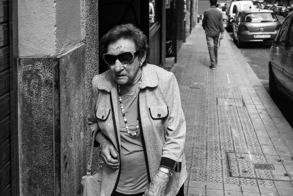 Ältere Frau mit Sonnenbrille geht auf einem Bürgersteig; Autos im Hintergrund.