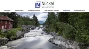 Altes rotes Gebäude am Flussufer, umgeben von Wald und Hügeln im Hintergrund.