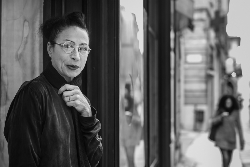 Frau mit Brille vor Schaufenster, Mantel tragend, unscharfe Person im Hintergrund, Schwarz-Weiß.