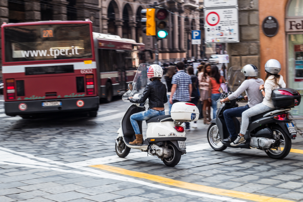 Mehrere Motorroller auf belebter Straße, roter Bus und Fußgänger im Hintergrund sichtbar.