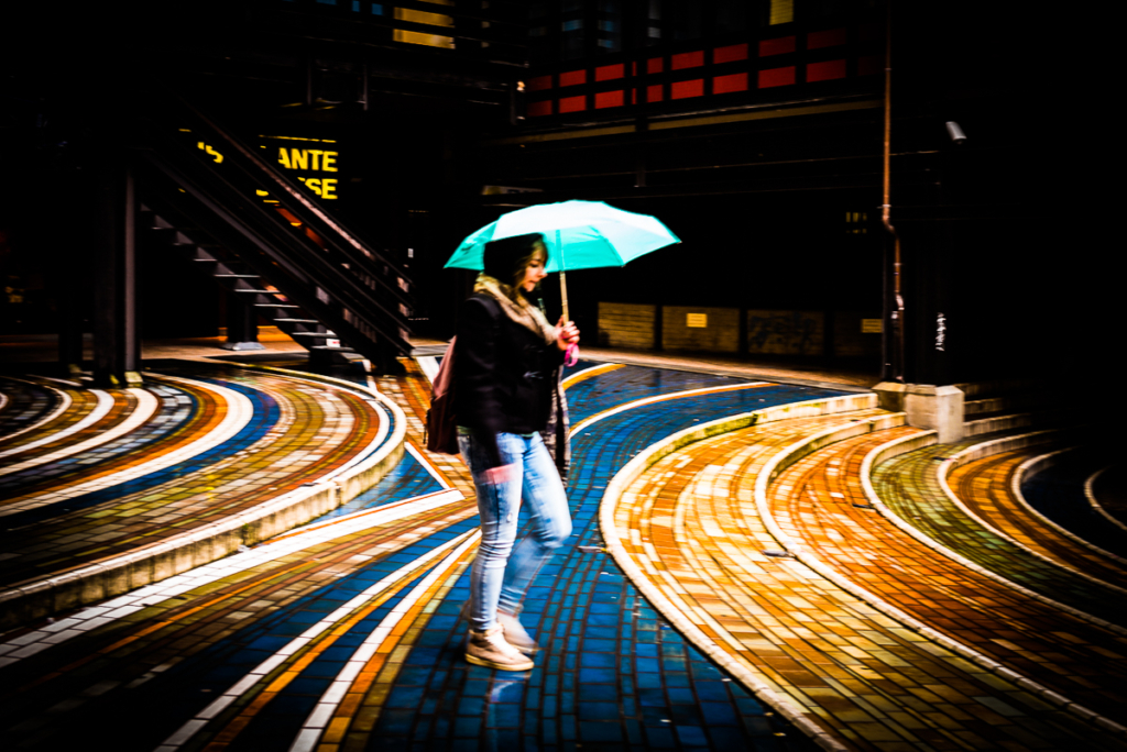 Person mit türkisfarbenem Regenschirm auf buntem, geschwungenem Boden in urbaner Umgebung.