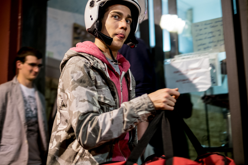 Person mit weißem Helm und Tarnjacke trägt Tasche vor einem Gebäude mit Glastüren.