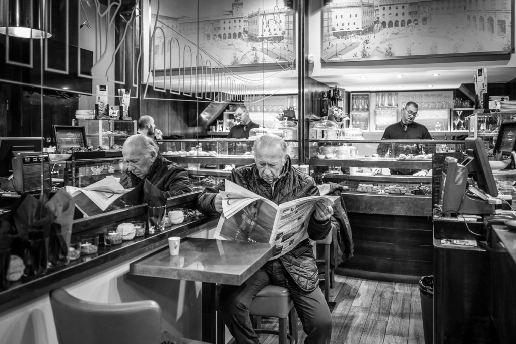 Älterer Mann liest Zeitung im Café, mit Tresen, Snacks und Bildern im Hintergrund.