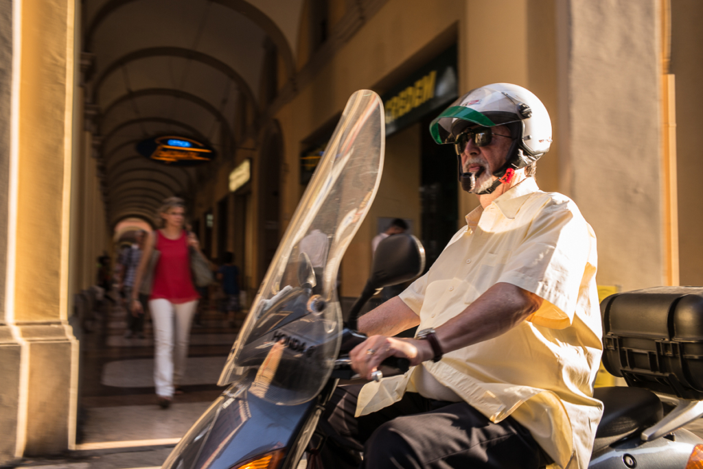 Mann mit Helm und Sonnenbrillen fährt Motorroller in überdachtem urbanem Bereich, Frau geht vorbei.