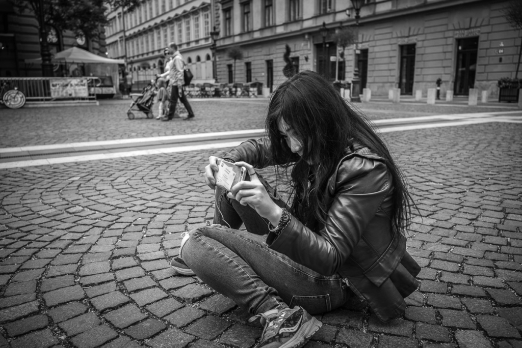 Person sitzt auf gepflastertem Platz mit Smartphone, Passanten im Hintergrund auf der Straße.