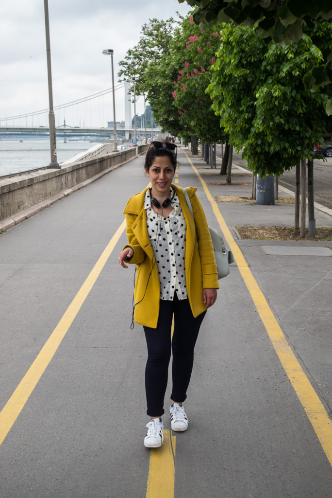 Person in gelbem Mantel spaziert am Fluss auf Gehweg, mit Bäumen und Brücke im Hintergrund.