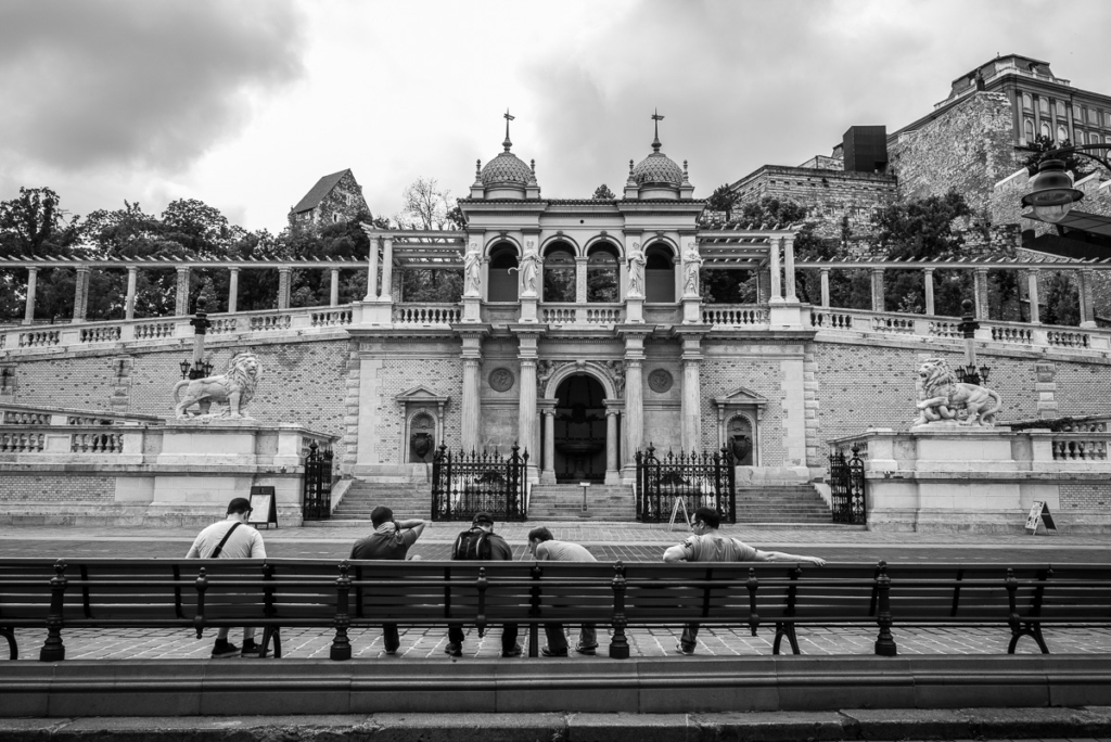 Menschen auf einer Bank vor historischer Gebäudefassade mit Säulen und Skulpturen.
