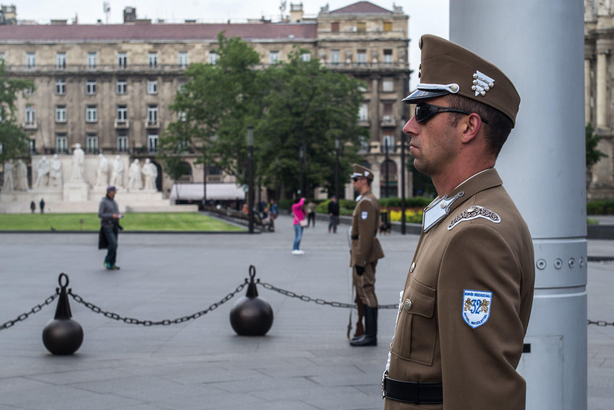 Soldat in brauner Uniform und Sonnenbrille vor städtischem Gebäudekomplex mit Statue und Passanten.