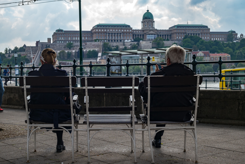 Zwei Personen sitzen auf Bänken mit Blick auf ein großes historisches Gebäude auf einem Hügel.