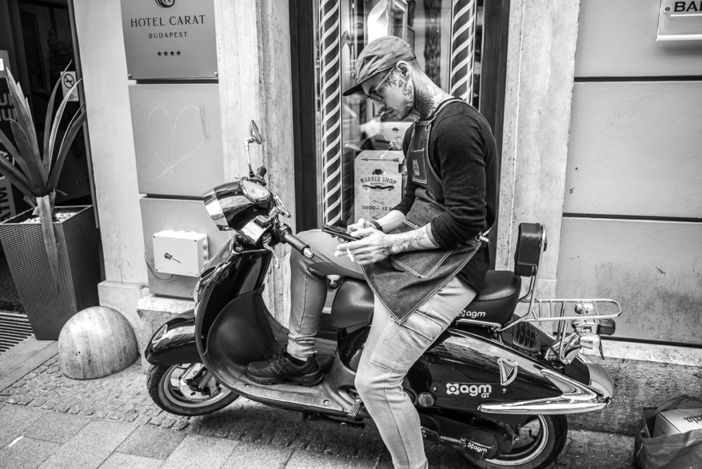 Mann auf Roller vor Hotel in Budapest, schaut auf Mobiltelefon auf gepflastertem Gehweg.