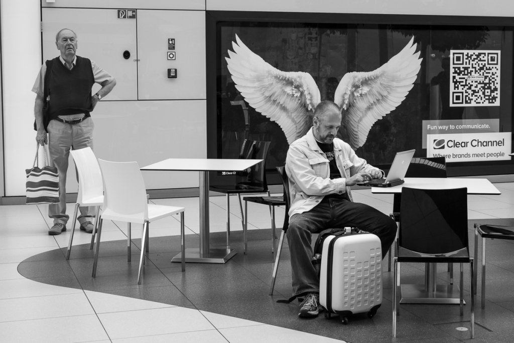 Mann mit Laptop am Tisch, Poster mit Engelsflügeln im Hintergrund, anderer Mann mit Tasche.