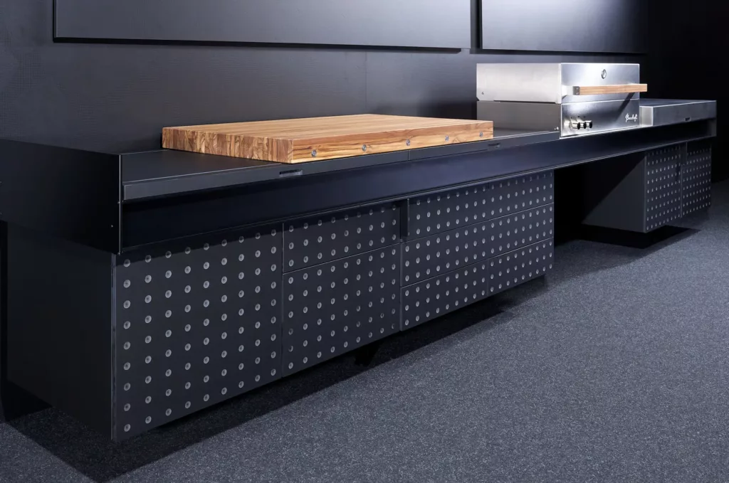 Modernes Küchenmöbel in Dunkel mit Schubladen und hellem Punktmuster, massives Holzbrett oben.