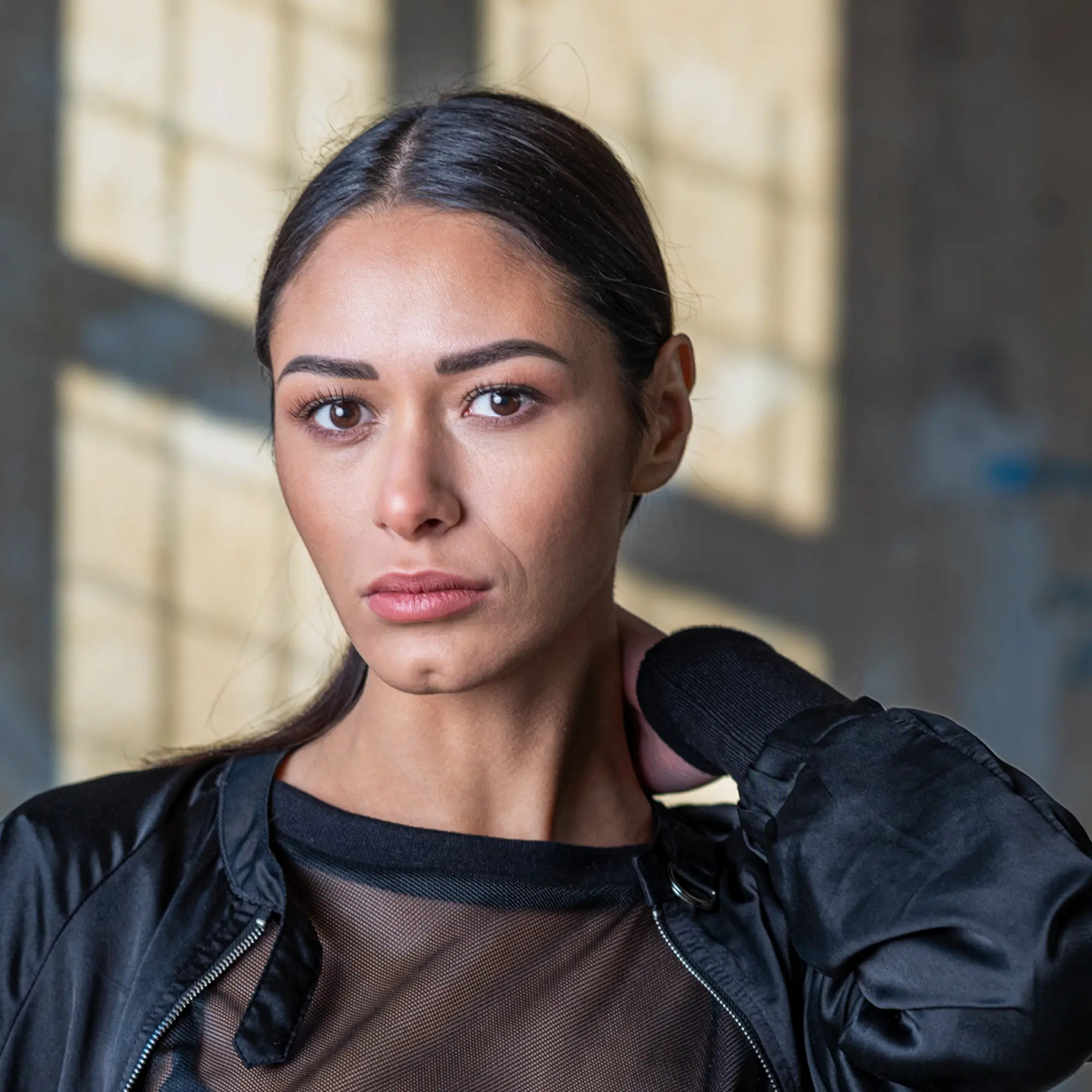 Frau mit dunklem Haar trägt schwarze Jacke vor unscharfem Hintergrund mit Fenster und Licht.