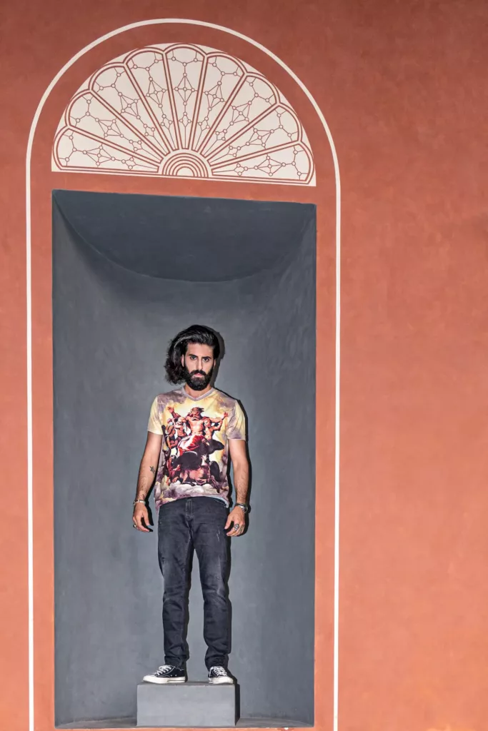 Mann in nischenartiger Öffnung vor verzierter Wand, trägt künstlerisches T-Shirt und sportliche Schuhe.