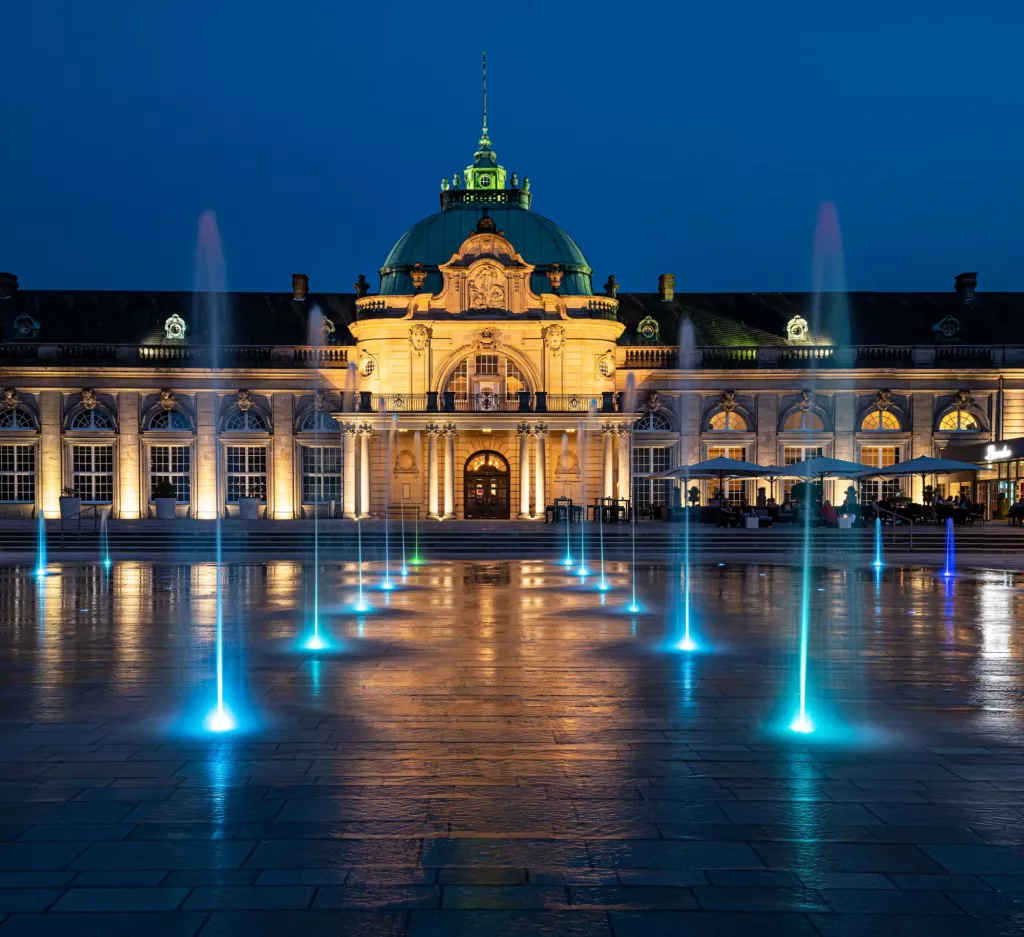 Historisches Gebäude bei Nacht, Kuppel, beleuchtete Springbrunnen auf gepflastertem Platz.