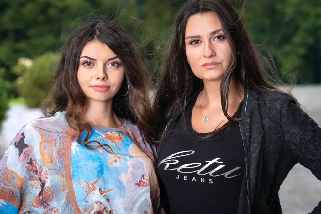 Zwei Frauen im Freien, bunte Kleidung links, schwarzes "Ketu Jeans" Oberteil rechts, Naturhintergrund.