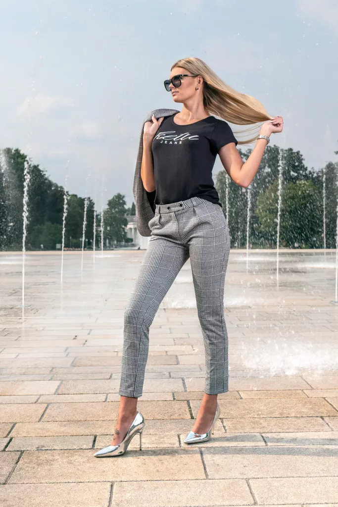Person vor Springbrunnen mit schwarzem T-Shirt, karierter Hose, silbernen High Heels und Sonnenbrille.