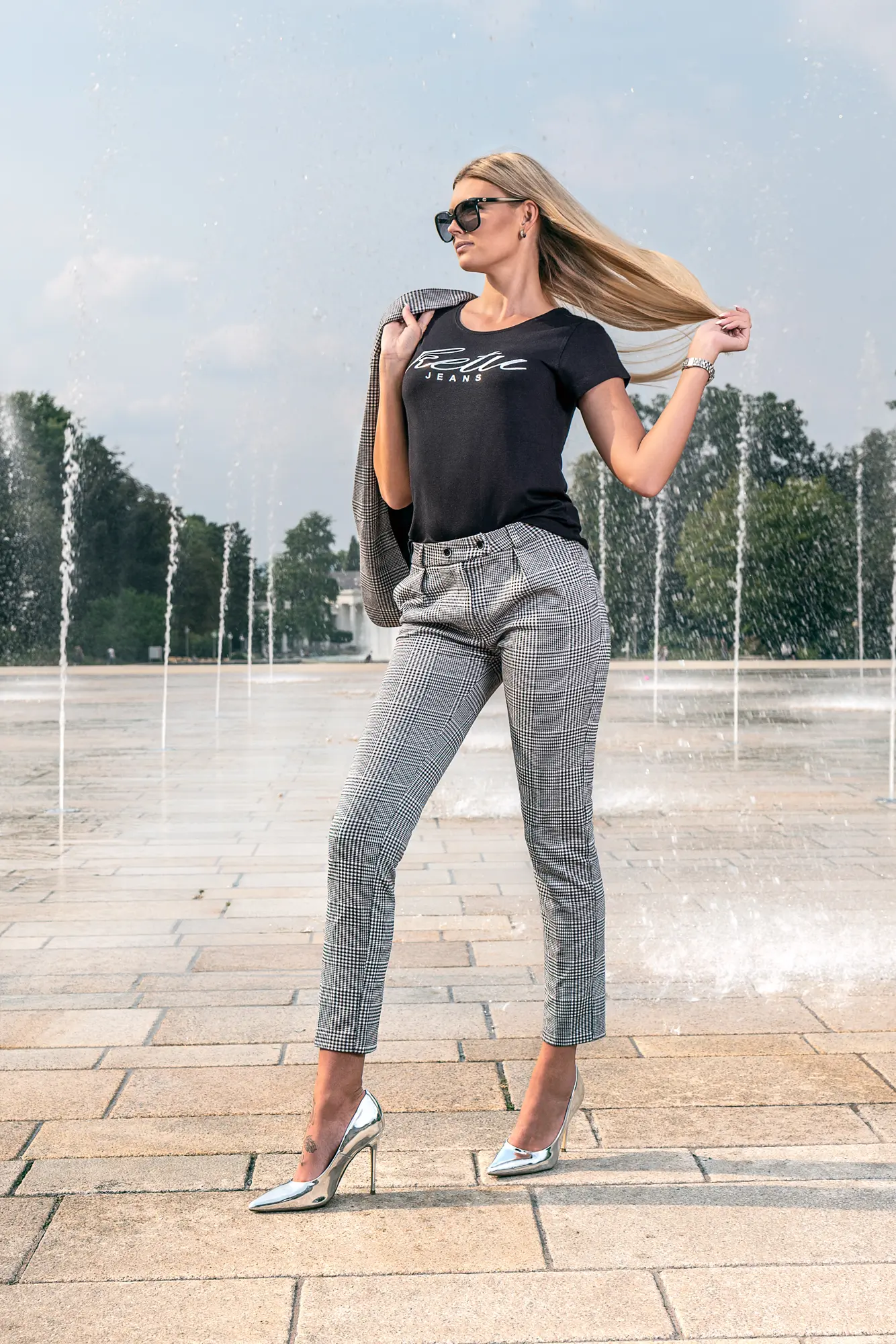 Person vor Springbrunnen mit schwarzem T-Shirt, karierter Hose, silbernen High Heels und Sonnenbrille.
