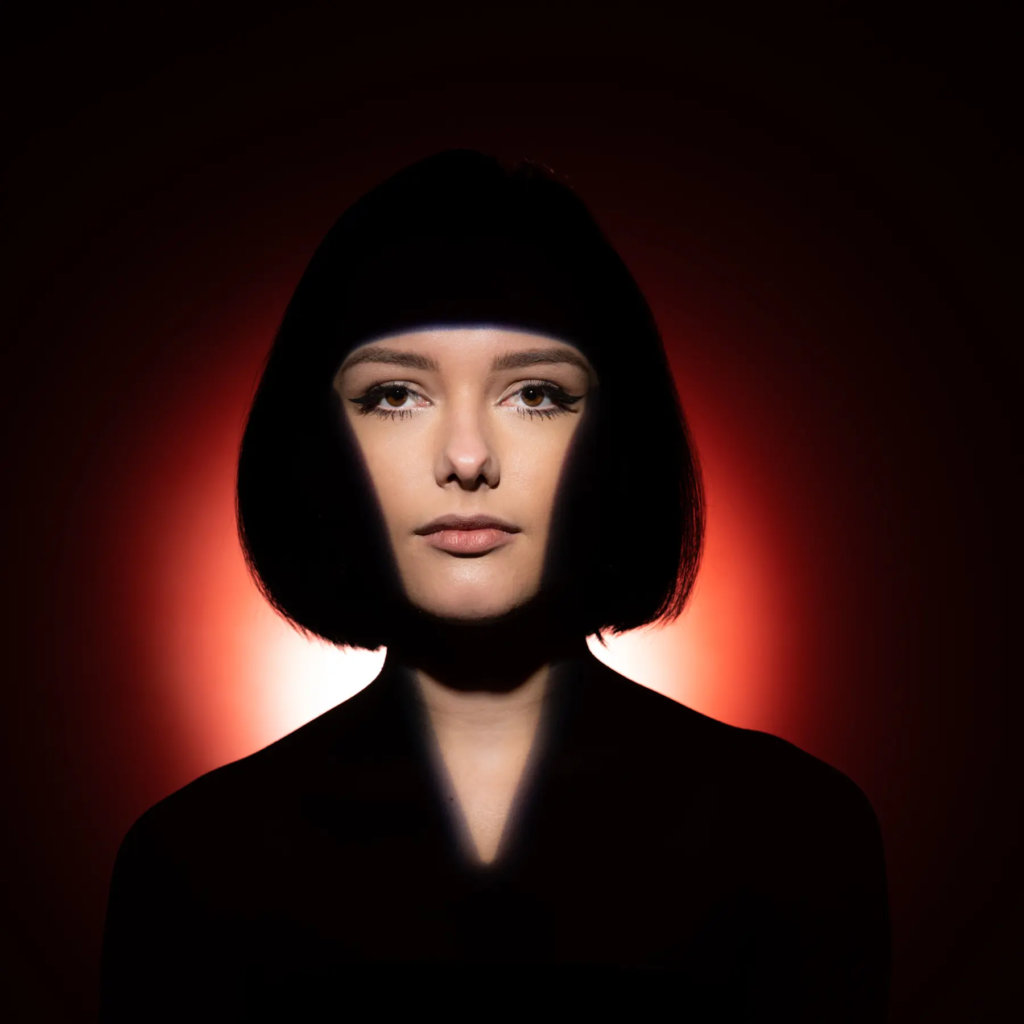 Frau mit kinnlangem Haar vor rotem Hintergrund, Gesicht teils von Schatten beleuchtet.