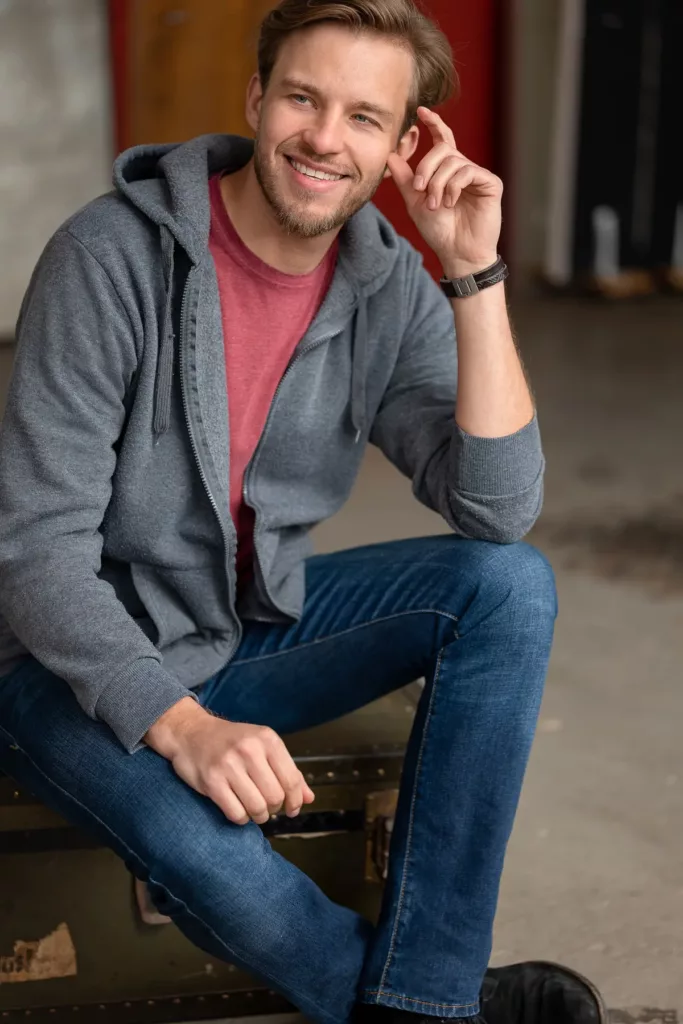 Mann in grauem Hoodie und Jeans sitzt lächelnd auf einem Koffer.