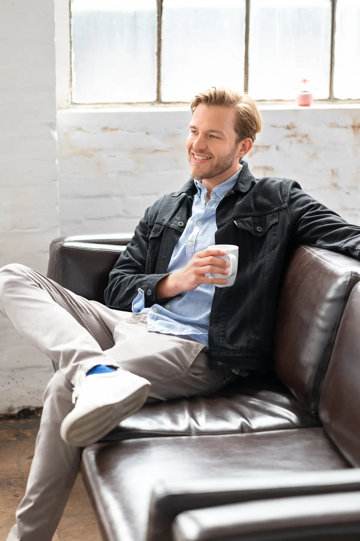 Person entspannt auf Ledersofa mit Tasse, trägt helle Hose, Hemd, Jacke, Fenster im Hintergrund.