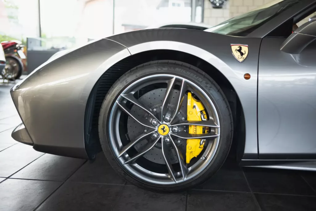 Grauer Sportwagen, linkes Vorderrad mit gelbem Bremssattel und Emblem, auf schwarzen Fliesen.